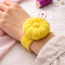 Yellow fabric pin cushion on a person's wrist with pins sticking out, on a light pink surface.