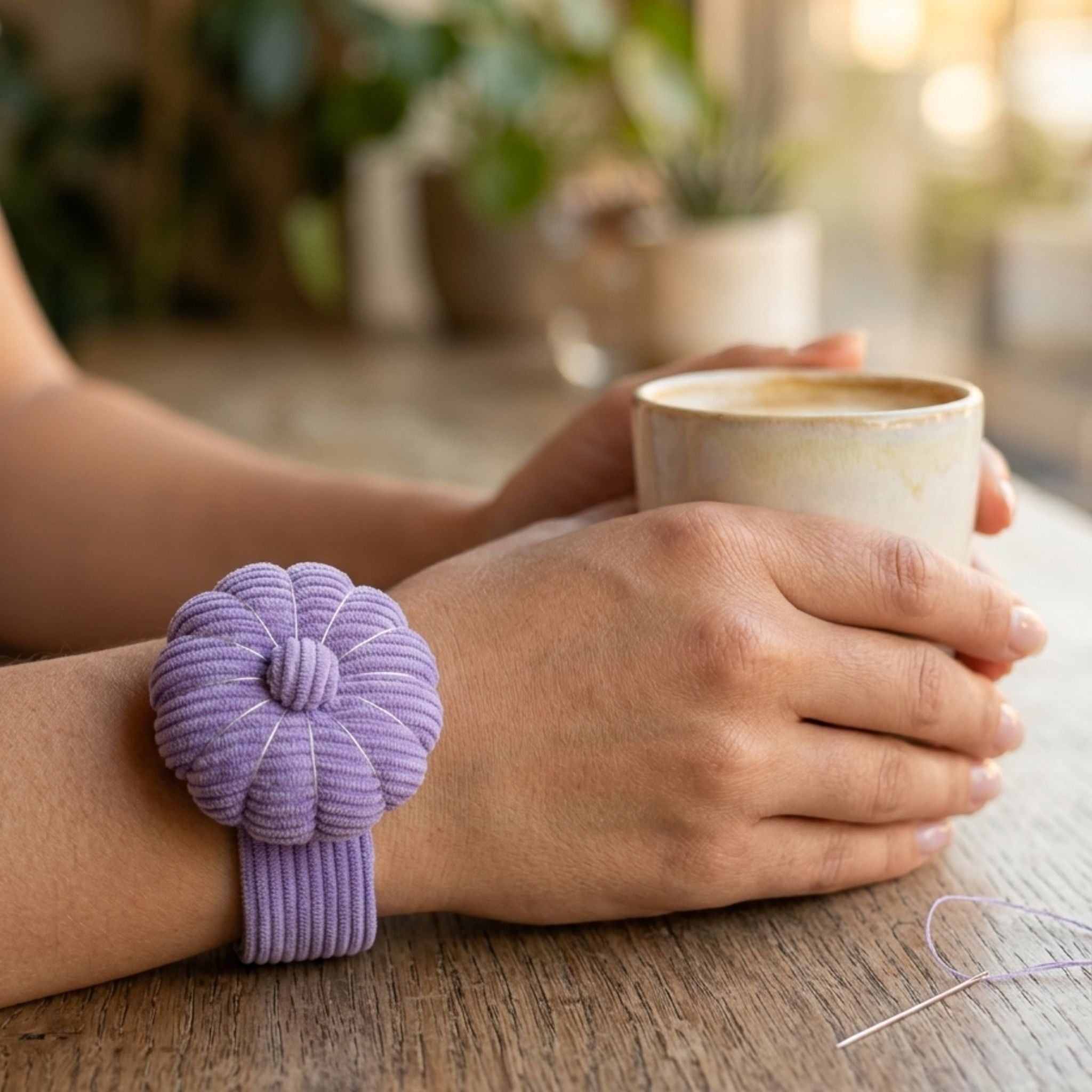 Person holding a cup wearing a Wild Lilac Pin Cushion on a wooden surface